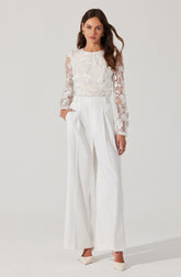 A woman is wearing a white long-sleeved blouse with lace floral patterns on the sleeves and front. She pairs it with high-waisted, wide-leg white trousers that feature pleats at the waist. She completes the outfit with white heels.